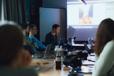 Participants in a workshop sit around a table and look at a projector screen displaying a presentation. | © Evolumina