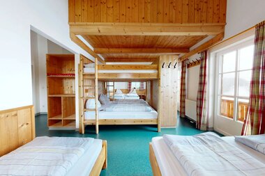 Cozy bedroom with wooden furniture and large windows overlooking the outdoors, in a rustic chalet or mountain cabin style. | © Pinzgauerhütte
