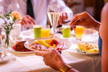 Table set with food, including a plate with cheese, ham, fruit, and jam. Drinks and flowers are visible in the background. | © Salzburgerhof Zell am See, Peter Kuehnl