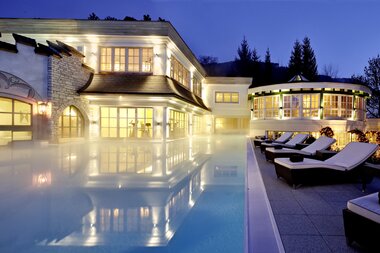 Elegant hotel in the evening with illuminated outdoor pool and loungers around the pool, surrounded by modern architecture. | © Salzburgerhof Zell am See