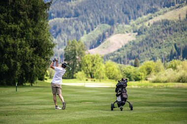 Golfer swings club on a lush green golf course with forested mountains in the background.