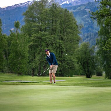 Golf player putting on a sunny green course surrounded by lush trees with mountains in the background.