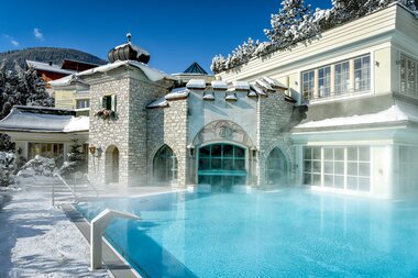 Winter scene of a hotel with an outdoor pool, surrounded by snow and a mix of traditional and modern buildings.