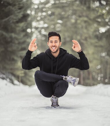 Person in a squat position in a snowy winter landscape, smiling with raised hands. | © Katharina Eder Arts 