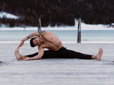 Person practicing yoga on a wooden platform in the mountains with water and snow in the background, stretching pose. | © Katharina Eder Arts 