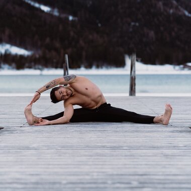 Person practicing yoga on a wooden platform in the mountains with water and snow in the background, stretching pose. | © Katharina Eder Arts 