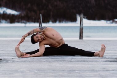 Person practicing yoga on a wooden platform in the mountains with water and snow in the background, stretching pose. | © Katharina Eder Arts 