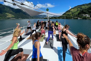 Yoga retreat on a boat with a lake and green mountains in the background, people practicing yoga outdoors. | © Zell am See-Kaprun Tourismus