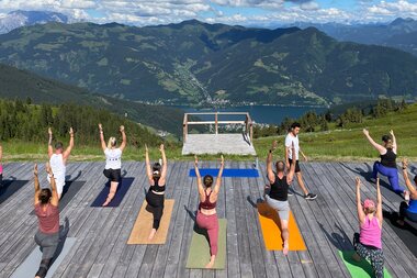 People practicing yoga on a wooden platform with mountain and lake view under a cloudy sky. | © Zell am See-Kaprun Tourismus