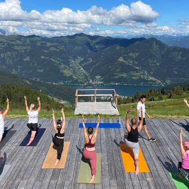People practicing yoga on a wooden platform with mountain and lake view under a cloudy sky. | © Zell am See-Kaprun Tourismus