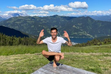 Person in Lotus position on a wooden platform, surrounded by green meadow and mountains under sunny sky. | © Zell am See-Kaprun Tourismus