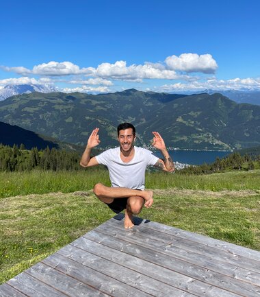 Person in Lotus position on a wooden platform, surrounded by green meadow and mountains under sunny sky. | © Zell am See-Kaprun Tourismus