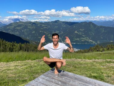 Person in Lotus position on a wooden platform, surrounded by green meadow and mountains under sunny sky. | © Zell am See-Kaprun Tourismus