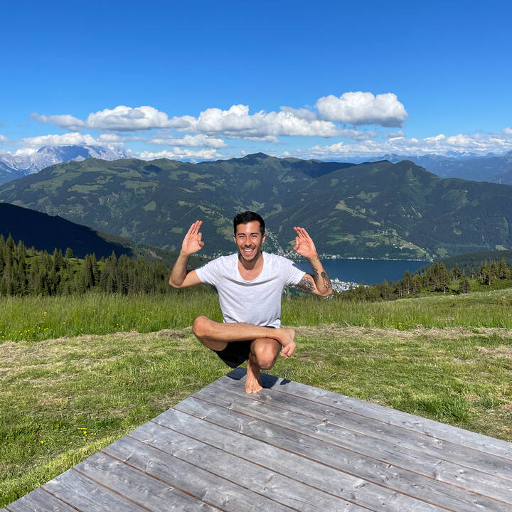 Person in Lotus position on a wooden platform, surrounded by green meadow and mountains under sunny sky. | © Zell am See-Kaprun Tourismus