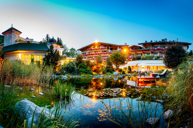 Evening view of a hotel with illuminated balconies, surrounded by a garden and water lilies on a pond. | © 5*S Hotel Salzburgerhof Zell am See