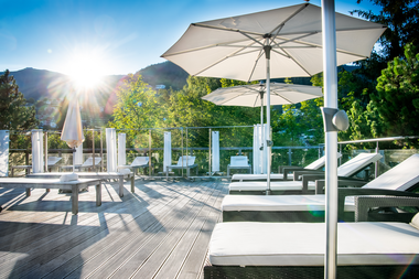 Sunny terrace with loungers, umbrellas, and a view of green trees and mountains during sunrise or sunset. | © 5*S Hotel Salzburgerhof Zell am See