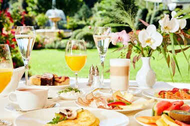 Breakfast table outdoors with a variety of foods such as fruit, tea, sparkling wine, and pastries in sunny weather. | © 5*S Hotel Salzburgerhof Zell am See