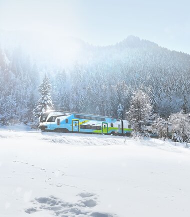 A train travels through a snowy winter landscape with trees and mountains in the background. | © Westbahn