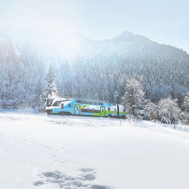 A train travels through a snowy winter landscape with trees and mountains in the background. | © Westbahn