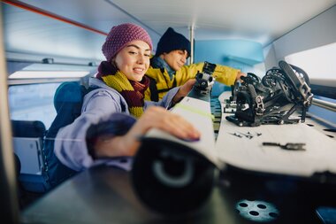 Two people in winter clothing on a train, one writing in a notebook, the other packing skis. | © Westbahn, Ulrich Zinell