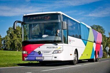 A modern colorful Postbus on a road with green landscape in the background. | © Postbus 