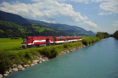 A red train travels along a river through a green landscape with mountains in the background. | © Salzburg AG