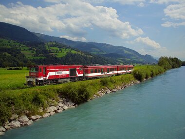 A red train travels along a river through a green landscape with mountains in the background. | © Salzburg AG