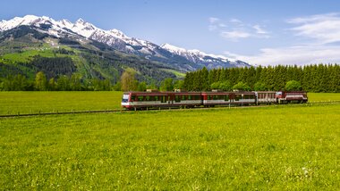 A train travels through a green meadow landscape with snow-capped mountains in the background under a clear blue sky. | © Zell am See-Kaprun Tourismus, Artisual