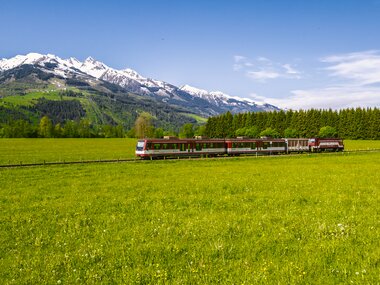 A train travels through a green meadow landscape with snow-capped mountains in the background under a clear blue sky. | © Zell am See-Kaprun Tourismus, Artisual
