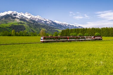 A train travels through a green meadow landscape with snow-capped mountains in the background under a clear blue sky. | © Zell am See-Kaprun Tourismus, Artisual