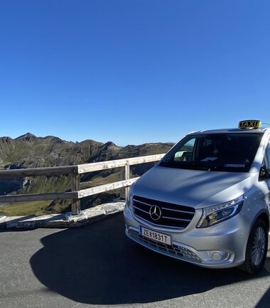 Silver Mercedes taxi in a mountainous landscape with blue sky and wooden railing | © Kapruner Taxi