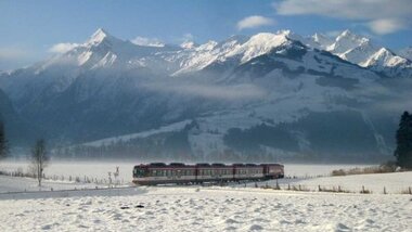 A train travels through a snowy winter landscape with tall, snow-covered mountains under a blue sky. | © Salzburg AG