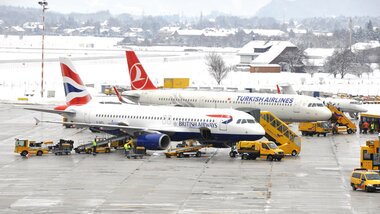 Two-plane at Salzburg Airport covered in snow, with more aircraft and airport buildings in the background. | © Salzburg Airport