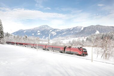 An ÖBB train traverses a snowy winter landscape with mountains in the background. | © ÖBB, Harald Eisenberger
