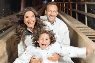 Family enjoying a slide in the Salt Mine Hallein, with a child smiling happily. | © Salzbergwerk Hallein 