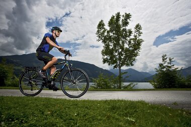 A person riding an e-bike along a path with water, trees, and mountains in the background. | © Zell am See-Kaprun Tourismus