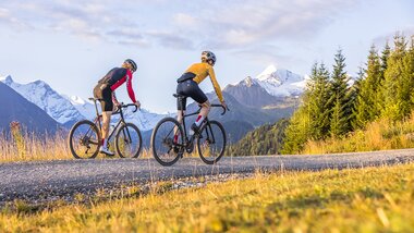 Two cyclists riding in a mountainous landscape with snow-capped peaks in the background during a gravel ride. | © SalzburgLand Tourismus