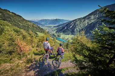 Two mountain bikers riding on a narrow trail with a view of a lake and mountains. | © Kitzsteinhorn