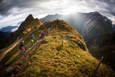 Mountain bikers on a narrow trail along a ridge in the Alps, surrounded by high mountains and clouds. | © David Schultheiss