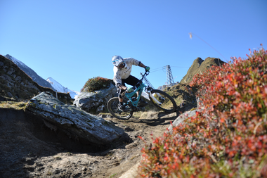 Mountain biker on a narrow trail in the mountains near Kitzsteinhorn, with colorful shrubbery in the foreground and mountain peaks in the background. | © Kitzsteinhorn 