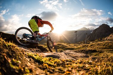 Mountain biker riding on a rocky trail in the mountains at sunset, with mountain and cloud scenery in the background. | © David Schulheiß 