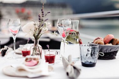 Elegant breakfast table set with flowers, wine glasses, water jug, and macarons with a water view. | © Birgit Schattbacher