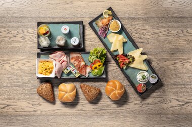 Breakfast platter with various bread types, cold cuts, cheese, eggs, and side dishes on a wooden table. | © Kitzsteinhorn 