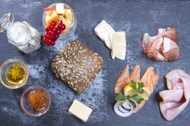 Various breakfast ingredients on a dark stone board, including cheese, cold cuts, olive oil, and fresh herbs. | © Kitzsteinhorn 