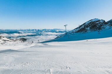 View of a snowy ski slope with skis in the foreground, surrounded by mountains and clear blue sky. | © Johannes Radlwimmer