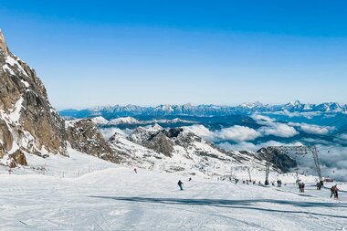 Snow-covered mountain slope under a clear sky with distant mountain peaks, skiers on the slope. | © Johannes Radlwimmer