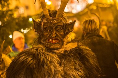 A person in a demon costume with horns and fur during a festive street event with warm lighting and decorations. | © Nikolaus Faistauer Photography