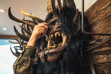 Person adjusting a large, mystical devil mask with horns on a wooden wall installation. | © Zell am See-Kaprun Tourismus