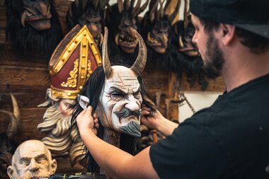 Artist crafting a demon mask with horns in a mask workshop, surrounded by other mask heads. | © Zell am See-Kaprun Tourismus