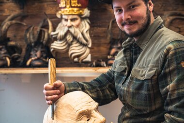 Artist working on a wooden mask sculpture in a workshop, surrounded by various masks on the wall. | © Zell am See-Kaprun Tourismus